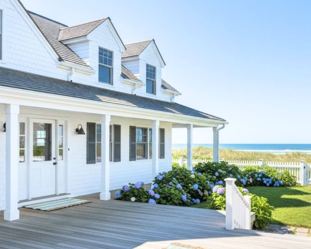 Charming white house with a front porch, blooming garden, and lush greenery under a clear blue sky, cozy countryside home exterior inspiration.