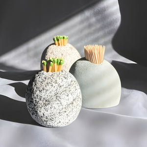 Three natural stones of varying shades - gray, tan, and charcoal - sitting on a gray fabric, two holding matches and one holding toothpicks.