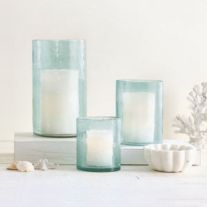 Blue hammered glass hurricane candle holders with candles on white tabletop with coastal decor