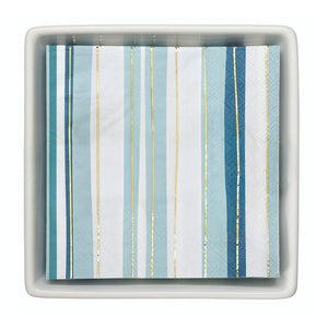 Blue striped cocktail napkins with gold foil accents displayed neatly inside a ceramic napkin tray