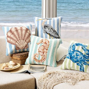 A collection of coastal-themed hook pillows, including a heron, seashell, and seahorse, arranged on a tan sofa overlooking a beach view.