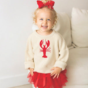 Toddler wearing chunky knit lobster sweater with red tutu skirt and red ribbon