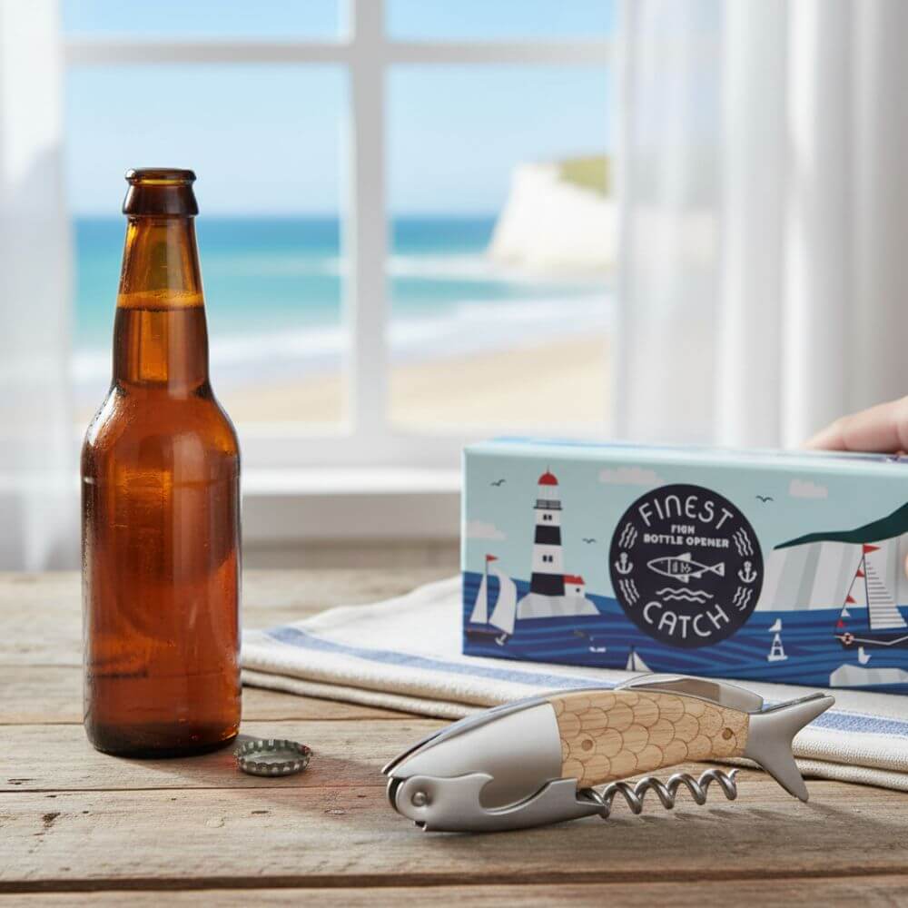 Fish-shaped bottle opener with wooden and stainless steel design on a wooden table by the beach, beside a beer bottle and nautical packaging.