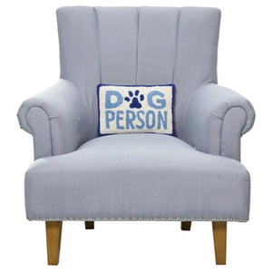 A blue "Dog Person" lumbar pillow centered on a light gray upholstered armchair with wooden legs.