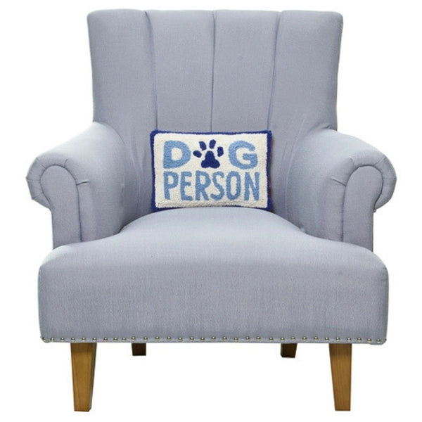 A blue "Dog Person" lumbar pillow centered on a light gray upholstered armchair with wooden legs.