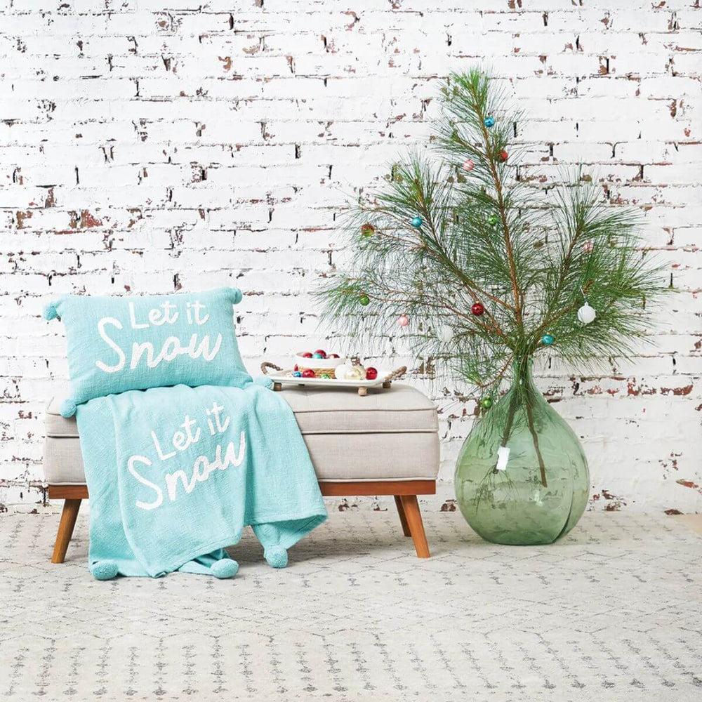Festive cotton pillow with pompom edging and winter-inspired “Let It Snow” design for seasonal décor.