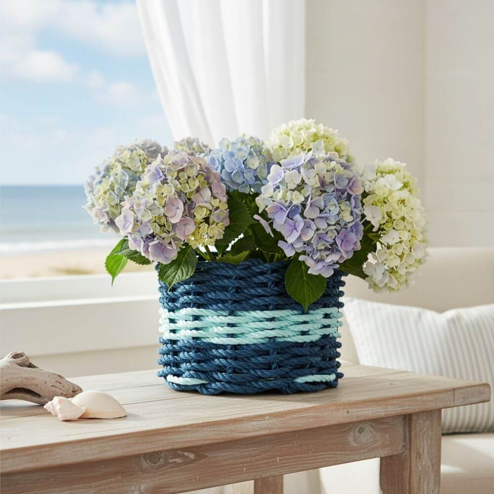 Beautiful bouquet of white and pale blue hydrangeas arranged in a blue and green woven basket, sitting on a rustic wooden surface with a serene beach and ocean view in the background, evoking a fresh coastal vibe.