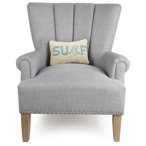 The cream and aqua "SURF" lumbar pillow resting in the center of a light grey upholstered wingback chair with silver nailhead trim.