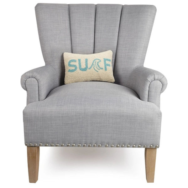 The cream and aqua "SURF" lumbar pillow resting in the center of a light grey upholstered wingback chair with silver nailhead trim.