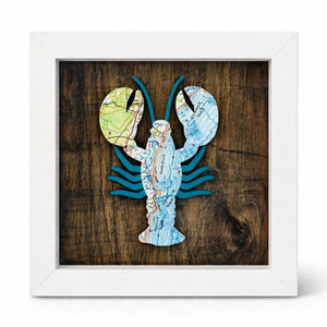 Framed artwork of a lobster made from nautical chart map pieces on a wooden background with a white wood frame