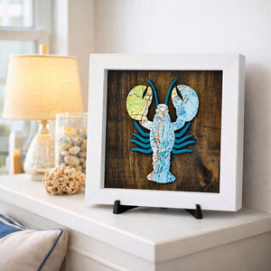 Framed artwork of a lobster made from nautical chart map pieces on a wooden background with a white wood frame on a side table