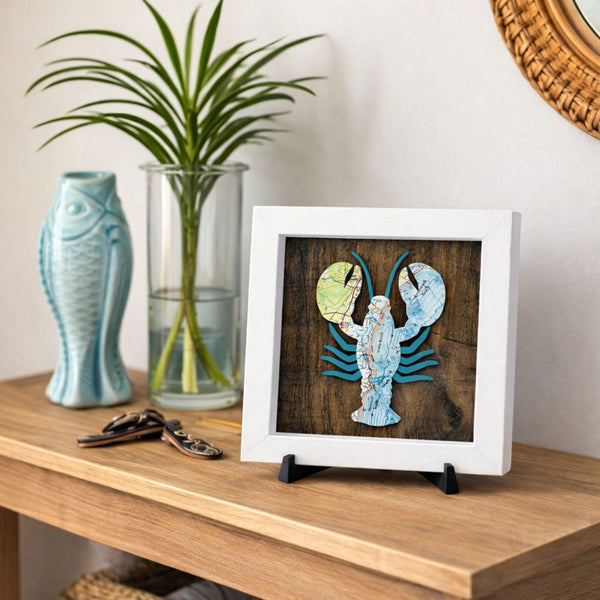 Framed artwork of a lobster made from nautical chart map pieces on a wooden background with a white wood frame on a table