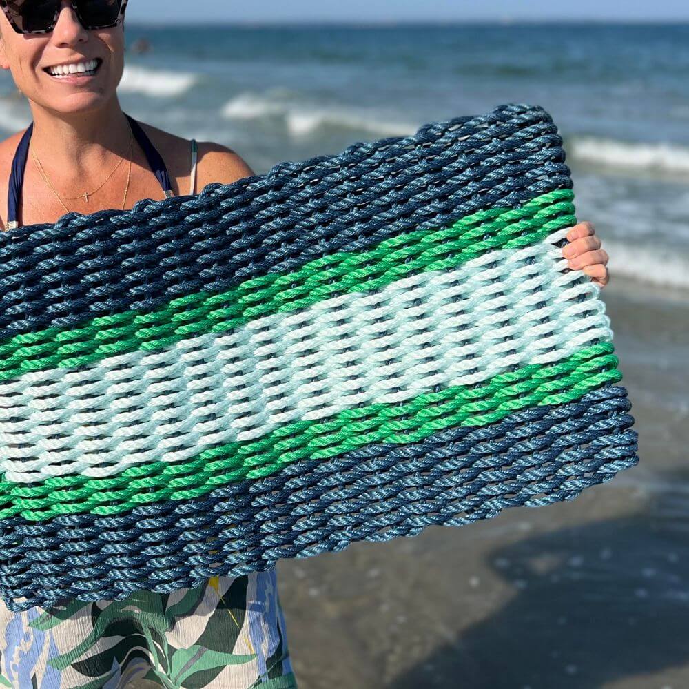 Person holding a large, intricately woven natural fiber mat with detailed patterns, standing outdoors with ocean waves crashing in the background under a clear sky.