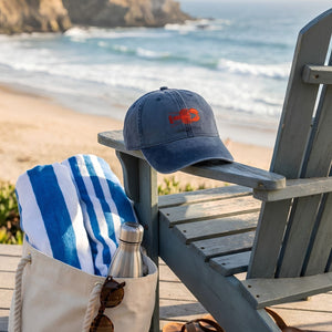 Washed Navy Baseball Hat With Red Lobster Embroidery Resting on a Weathered Beach Chair Beside a Blue and White Striped Beach Towel, Nautical Summer Accessory